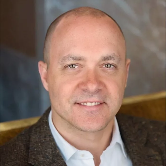 A bald man wearing a brown textured blazer and white shirt is seated indoors. The background features a neutral-toned wall and a golden chair. The setting suggests a formal or professional environment.